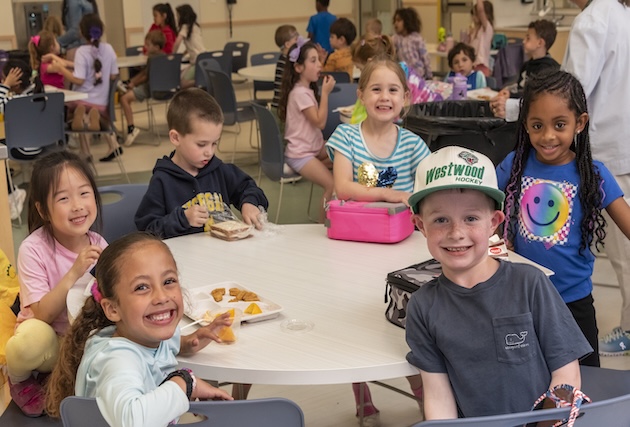 A group of students sit at a cafeteria table and smile for the camera.