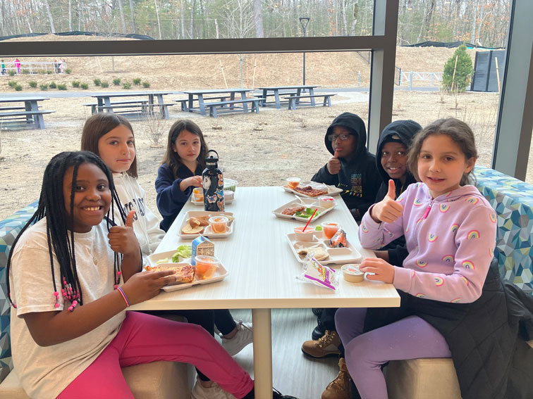 Six students sitting at a lunch table in front of a large window