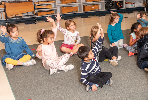 A classroom of students sitting on the floor with arms raised